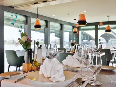 Elegantes Restaurant an Bord der MS Alisa mit gedeckten Tischen, Weingläsern und frischen Blumen. Große Fensterfront mit Blick auf den Fluss. Warme Pendelleuchten erhellen den Raum.