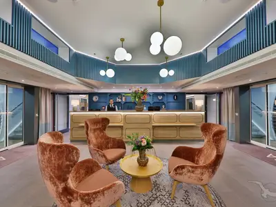 Modernes Foyer der MS Alisa. Helle Holzrezeption, ein Mitarbeiter. Vier samtige Sessel und ein Tisch im Wartebereich. Blaue Lamellen und auffällige Hängelampen.