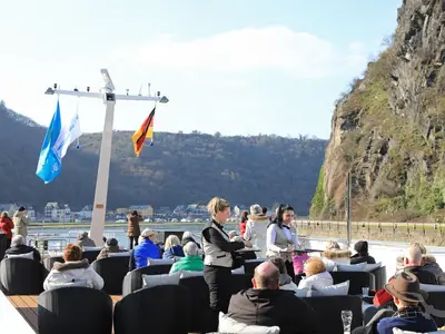Auf dem Sonnendeck der MS Alisa serviert Crew Getränke an Passagiere, die auf bequemen Korbsesseln sitzen. Im Hintergrund sind grüne Berge und ein Fluss zu sehen. Eine deutsche Flagge weht an einem Mast.