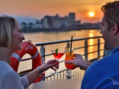 Ein Mann und eine Frau stoßen auf dem Sonnendeck der MS Alisa mit Cocktails an, während die Sonne über dem Fluss untergeht. Im Hintergrund ist eine Stadtkulisse zu sehen.