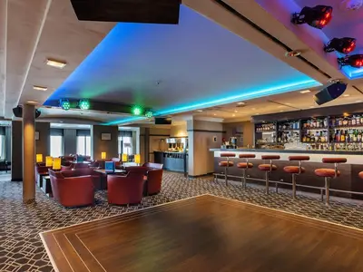 Die moderne Harry's Bar an Bord der MS Amadea mit einer gut bestückten Theke, Barhockern, roten Sesseln und Tischen. Die Decke ist mit blauen und grünen LED-Leuchten illuminiert, davor eine Tanzfläche aus Holz.
