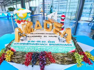 Ein festlicher, mehrstöckiger Kuchen mit der Aufschrift 'AMADEA' und 'WILLKOMMEN AN BORD' für die MS Amadea. Er ist maritim dekoriert mit Ankern, Steuerrädern, einem Leuchtturm und einer Segeljacht. Um den Kuchen liegen Trauben. Im Hintergrund ist eine moderne Halle mit großen Fenstern zu sehen.