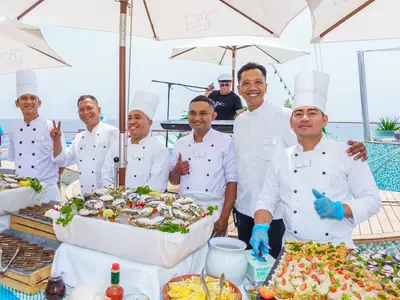 Sechs lächelnde Köche in Uniform präsentieren Austern und Desserts auf einem Buffet an Bord der MS Amadea. Im Hintergrund ein Musiker auf der Bühne und das Meer.