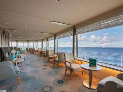 Blick in die helle Vista Lounge an Bord der MS Amadea. Gemütliche Sitzgruppen und große Fenster mit Panoramablick auf das Meer, am Horizont ein Schiff.
