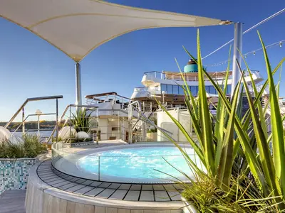 Blick auf einen runden Whirlpool mit klarem Wasser auf dem Teakholz-Sonnendeck der MS Amadea. Pflanzen in mosaikbesetzten Beeten und das Schiffsgebäude im Hintergrund sind sichtbar.