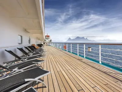 Promenadendeck der MS Amadea mit leeren Liegestühlen. Der Blick geht über das Geländer auf das weite, blaue Meer und eine ferne Bergkette unter bewölktem Himmel.