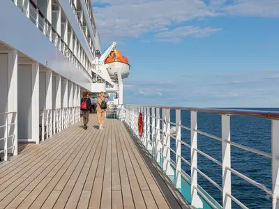 Zwei Frauen spazieren auf dem Holzdeck der Rundumpromenade der MS Amadea. Links sind Kabinenfenster, rechts das weite Meer unter blauem Himmel. Ein Rettungsboot hängt an Backbord.