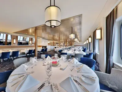 Geräumiger Speisesaal des Restaurants Vier Jahreszeiten auf der MS Amadea. Viele festlich gedeckte Tische, blaue und graue Stühle. Elegante Beleuchtung durch Fenster und Lampen.