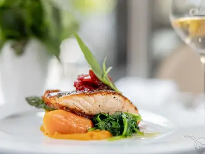 Detailaufnahme eines köstlich angerichteten Lachsfilets mit Spinat, Karottenpüree und roter Salsa. Dazu ein Weinglas, serviert an Bord der MS Amadea.