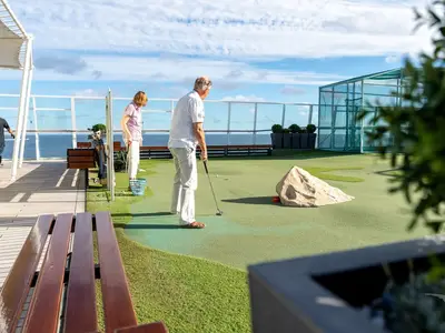 Ein Mann puttet auf einer Minigolfanlage auf dem Sonnendeck der MS Amadea. Eine Frau wartet am nächsten Loch. Im Hintergrund ist das blaue Meer zu sehen.