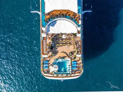 Vogelperspektive auf das Heckdeck der MS Amadea. Sichtbar sind mehrere Pools, Sonnendecks mit blauen Liegen, Tische mit gelben Stühlen und Gäste. Das Schiff bewegt sich auf dem offenen Meer.
