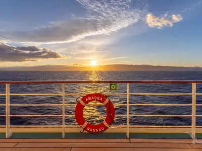 Atemberaubender Sonnenuntergang über dem weiten Meer, vom Deck der MS Amadea. Roter Rettungsring mit Schiffsnamen am Geländer. Berge am Horizont.