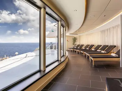 Eleganter Ruheraum an Bord der MS Amadea mit dunklen Liegestühlen und aufgerollten Handtüchern. Große Panoramafenster bieten einen weiten Blick auf das blaue Meer und den Himmel.