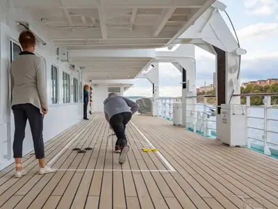 Ein Mann spielt Shuffleboard auf dem Teakdeck der MS Amadea, beobachtet von zwei Frauen. Im Hintergrund sind Gebäude an der Küste zu sehen.