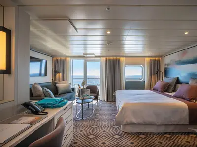 Geräumige Suite an Bord der MS Amadea mit großem Doppelbett, Sitzbereich mit Sofa und Beistelltisch, Schreibtisch, und Balkon mit Meerblick. Helles, modernes Design.