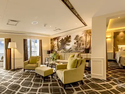 Geräumiger Wohnbereich einer Royal-Suite an Bord der MS Amadea. Beige Möbel, ein Dschungel-Wandbild und Meerblick durch die Fenster. Ein offener Durchgang führt zum Schlafzimmer.