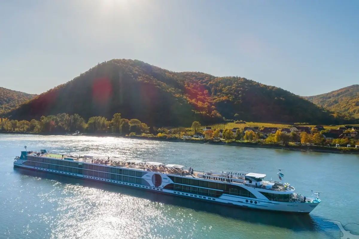 Luftaufnahme der MS Amelia auf einem Fluss im Herbst. Im Hintergrund bewaldete Berge mit Herbstlaub und ein Dorf mit Weinbergen. Sonnenlicht spiegelt sich auf dem Wasser.