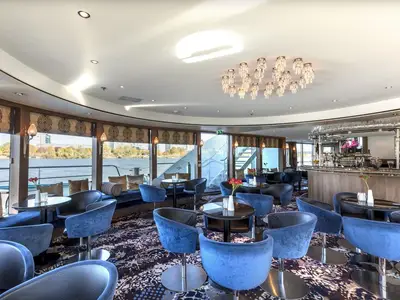 Die stilvolle Bar-Lounge der MS Amelia bietet blaue Samtstühle, dunkle Tische und eine Holztheke. Große Fenster erlauben einen Panorama-Blick auf einen Fluss und bewaldetes Ufer.