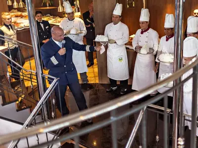 An Bord der MS Amelia präsentiert der Kapitän mit Mikrofon festliche Kuchen, die von mehreren Köchen getragen werden. Eine festliche Veranstaltung im Innenbereich des Schiffes mit weiteren Crewmitgliedern im Hintergrund.