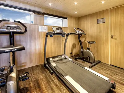 Innenansicht des Fitnessstudios an Bord der MS Amelia. Es sind ein Laufband und ein Ergometer zu sehen. Die Wände sind holzvertäfelt, mit Fenstern, die den Himmel zeigen. Der Raum ist menschenleer.