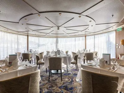 Ein elegantes Restaurant an Bord der MS Amelia mit weiß gedeckten Tischen und modernen Stühlen. Große Panoramafenster mit Vorhängen. Stimmungsvolle Deckenbeleuchtung.
