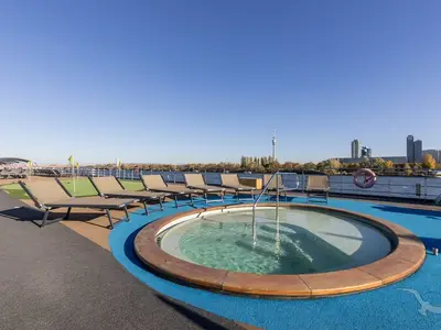 Sonnendeck der MS Amelia mit Whirlpool, Liegestühlen und Putting Green. Im Hintergrund ein Fluss und eine Stadt-Skyline mit Hochhäusern und einem Fernsehturm bei Tag.