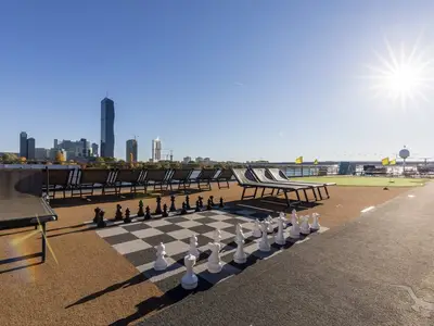 Das sonnige Oberdeck der MS Amelia zeigt ein großes Schachbrett, eine Minigolfanlage mit gelben Fahnen und Liegestühle. Im Hintergrund eine moderne Stadt-Skyline unter blauem Himmel.