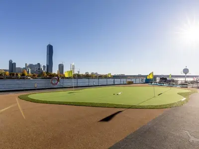 Ein sonniges Deck der MS Amelia mit grünem Mini-Golf-Putting-Green und gelben Flaggen. Im Hintergrund ein Fluss und eine moderne Stadt-Skyline unter blauem Himmel.