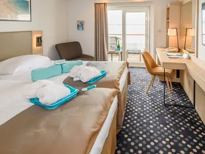 Moderne Balkonkabine an Bord der MS Amera mit zwei Betten, Schreibtisch, Sessel und Zugang zum Balkon. Helle Einrichtung, Bademäntel liegen bereit.