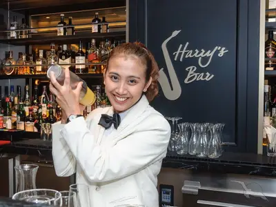 Eine lächelnde Barkeeperin mit schwarzer Fliege mixt einen Cocktail in Harry's Bar an Bord der MS Amera. Im Hintergrund sind Spirituosen und Gläser zu sehen.