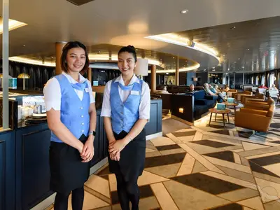 Zwei lächelnde Crew-Mitarbeiterinnen in blauen Westen stehen vor der Bar von Harry's Bar an Bord der MS Amera. Im Hintergrund entspannen Gäste in der modernen Lounge.