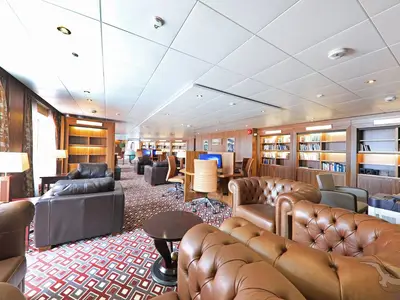 Innenansicht der Bordbibliothek der MS Amera mit zahlreichen Büchern in Holzregalen, bequemen braunen Ledersofas und -sesseln, sowie mehreren Computertischen für Gäste.