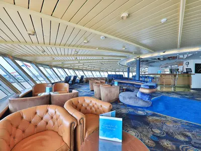 Die Panorama Lounge der MS Amera zeigt große Fenster mit Meerblick, bequeme braune und blaue Sitzgruppen sowie eine Bar.