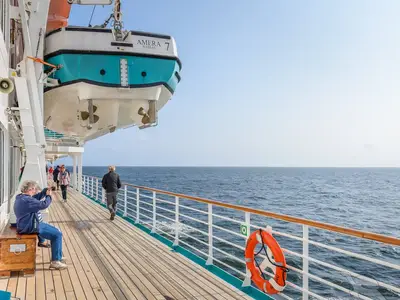 An Bord der MS Amera spazieren Gäste auf dem Teakholz-Promenadendeck. Links eine Frau mit Kamera, rechts ein Rettungsboot, daneben das offene Meer.
