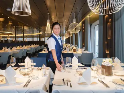 Lächeln. Eine Crewmitarbeiterin in blauer Uniform deckt einen Tisch im Restaurant Ozean der MS Amera. Viele elegant gedeckte Tische und eine helle Fensterfront prägen den modernen Raum.