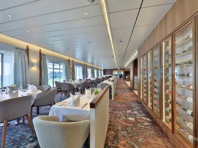 Blick in das stilvolle Restaurant Amera auf der MS Amera. Mehrere Tische sind weiß gedeckt mit Servietten und Gläsern, die Stühle sind grau. An der rechten Wand befindet sich eine lange, beleuchtete Weinvitrine mit vielen Flaschen. Durch große Fenster auf der linken Seite ist eine Außenansicht zu sehen.
