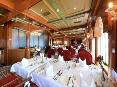 Spezialitätenrestaurant 'Pichler's' der MS Amera. Elegantes Holzinterieur, weißgedeckte Tische mit roten Samtstühlen, Gläsern und roten Blumen. Klassische Säulen und dekorierte Decken prägen die Atmosphäre.