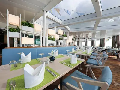 Helles Lido Wintergarten Restaurant auf der MS Amera. Gedeckte Tische mit grünen Platzsets, blauen Sitzbänken und einer Trennwand mit Pflanzen und Lichtern. Große Fenster.