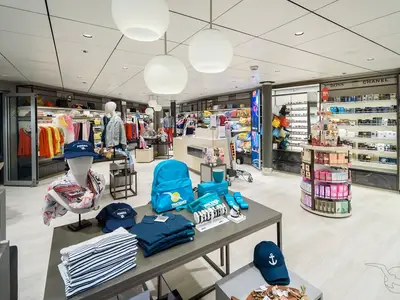 Ein weitläufiger, moderner Shop an Bord der MS Amera mit Kleidung, Taschen und Accessoires auf Tischen und Regalen. Links Kleidung, rechts Parfümtheke mit BOSS und CHANEL.