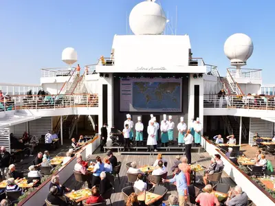Blick auf das obere Sonnendeck der MS Amera: Zahlreiche Gäste sitzen an Tischen. Auf einer Bühne stehen Köche vor einer großen Leinwand, die eine Weltkarte zeigt. Sonniges Wetter.