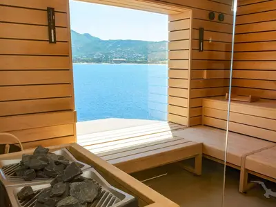 Sauna an Bord der MS Amera. Helle Holzeinrichtung, Bänke, Ofen mit Steinen. Durch die Glasfront blickt man auf weites, blaues Meer und grüne Berge.