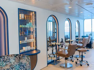 Heller, moderner Friseursalon an Bord der MS Amera mit braunen Lederstühlen, Spiegeln, Produktvitrine und Blick auf Meer und Berge. Ohne Personen.