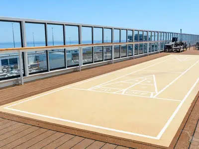 Ein leerer Shuffleboard-Platz mit Spielmarkierungen und Zahlen auf dem Deck der MS Amera. Im Hintergrund gläserne Geländer mit Blick auf das Meer und einen Hafenbereich.