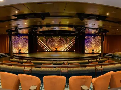 Ein weiter Blick in das leere Theater an Bord der MS Amera. Die Bühne ist mit goldenen und violetten Herzmustern beleuchtet, umgeben von orangefarbenen Sitzen und dunkler Atmosphäre.