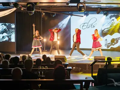 Vier Tänzer in Rock'n'Roll-Outfits treten auf einer Bühne im Theater der MS Amera auf. Im Hintergrund sind Bildschirme mit Elvis Presley und dem Schriftzug "Elvis" zu sehen, während das Publikum zuschaut.