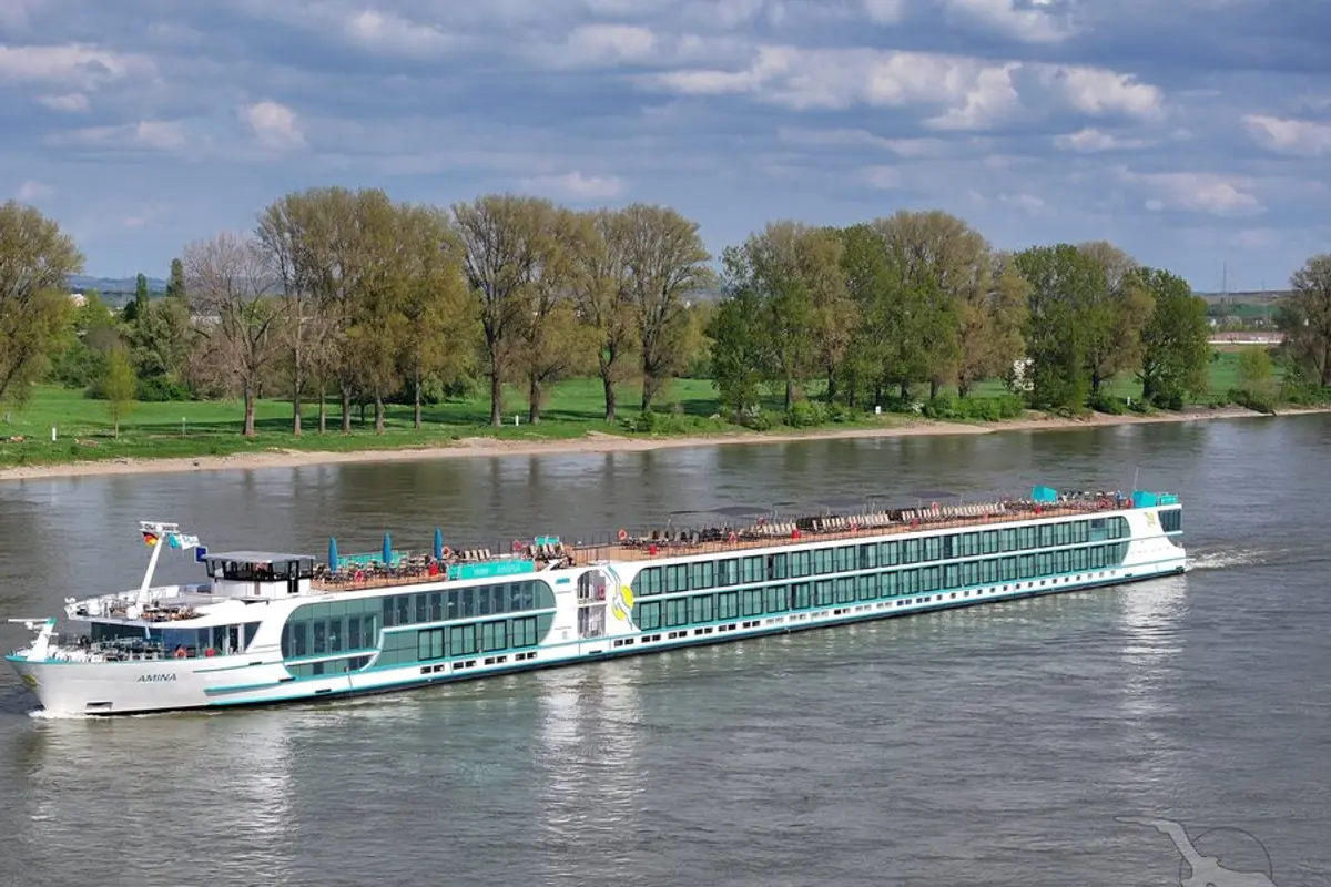 Flusskreuzfahrtschiff MS Amina auf einem breiten Fluss. Weiße Außenansicht mit türkisen Akzenten, vielen Fenstern und Sonnendeck. Grüne Bäume und Ufer am Horizont.
