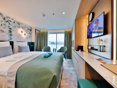 Eine modern eingerichtete Balkonkabine an Bord der MS Amina mit Doppelbett, Sitzbereich und großem Fenster zum Fluss. Die hellen Farben und floralen Akzente schaffen eine entspannte Atmosphäre.