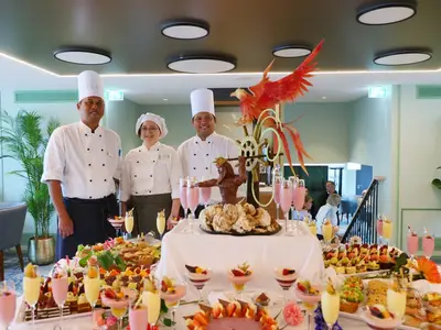 Drei lächelnde Crewmitglieder der MS Amina stehen hinter einem Buffet mit vielen Desserts, pinken Getränken und einer großen Schokoladenskulptur. Gäste sind im Hintergrund.