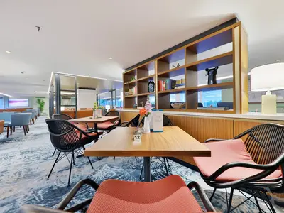 Eine moderne Panorama-Lounge an Bord der MS Amina mit Tischen, Rattan-Stühlen mit orangefarbenen Kissen, einem Bücherregal und Blick nach draußen. Der Boden ist mit einem blauen, gemusterten Teppich ausgelegt. Keine Personen sichtbar.
