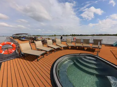 Das Sonnendeck der MS Amina mit braunen Liegestühlen und einem kleinen runden Pool. Ein Fluss und Uferbebauung sind im Hintergrund zu sehen. Wolken am blauen Himmel.